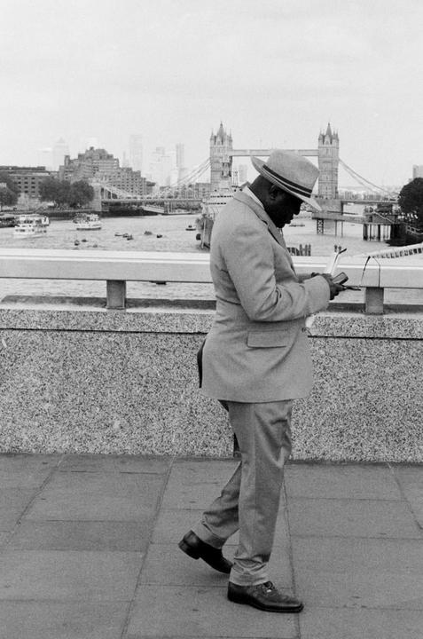 A man in a suit and hat walks along a bridge with the Tower Bridge in the background.
