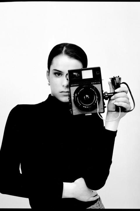 A woman in a black turtleneck holds a camera up to her face, obscuring one eye.