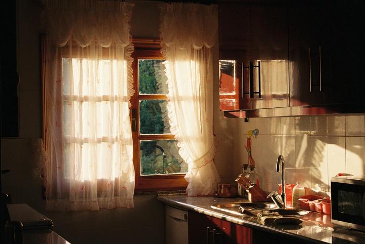 Sunlight streams through a window with sheer curtains into a kitchen with a sink and microwave.