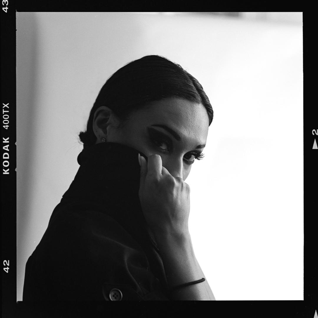 A woman with dark eye makeup looks over her shoulder while holding her collar up to her face.