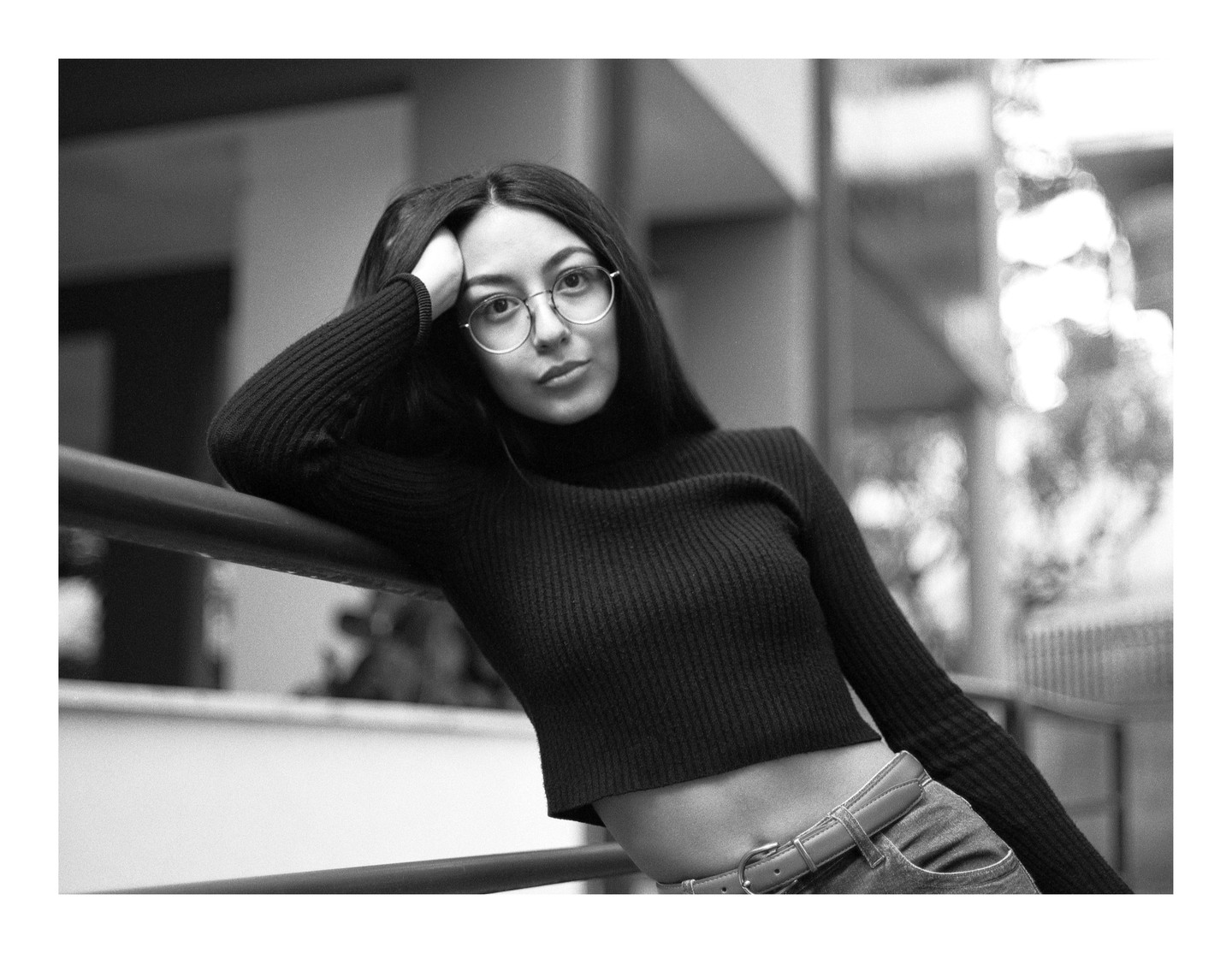 A woman with long hair and glasses leans on a railing, looking directly at the viewer.