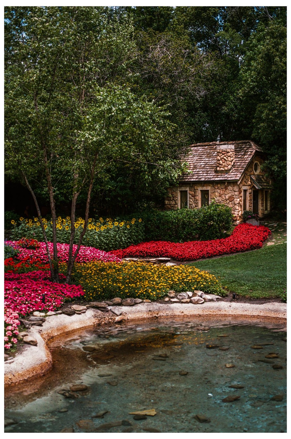 A small stone cottage with a wooden roof is nestled among trees and colorful flowerbeds next to a pond.