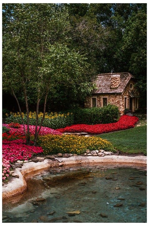 A small stone cottage with a wooden roof is nestled among trees and colorful flowerbeds next to a pond.