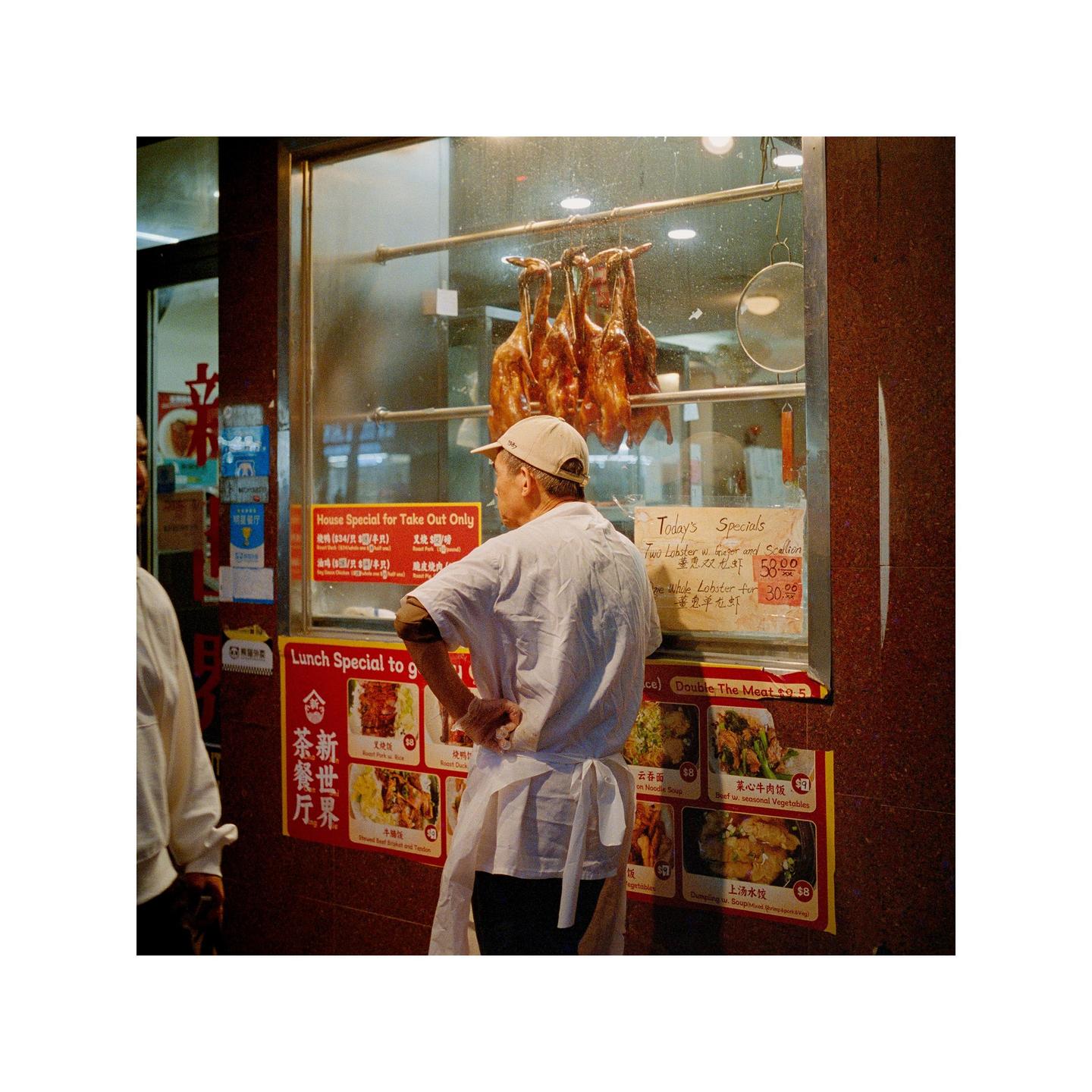 A man wearing a cap and apron stands in front of a restaurant window displaying roasted ducks and various menu items.