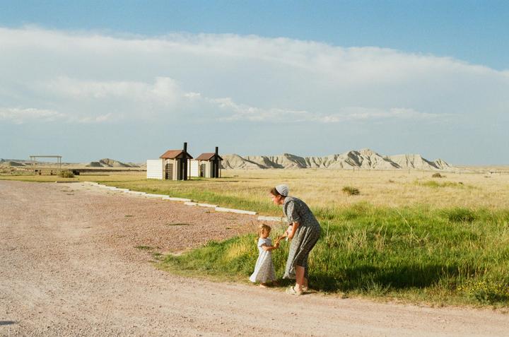 A woman and a child stand on a dirt road next to a grassy field with mountains in the distance.