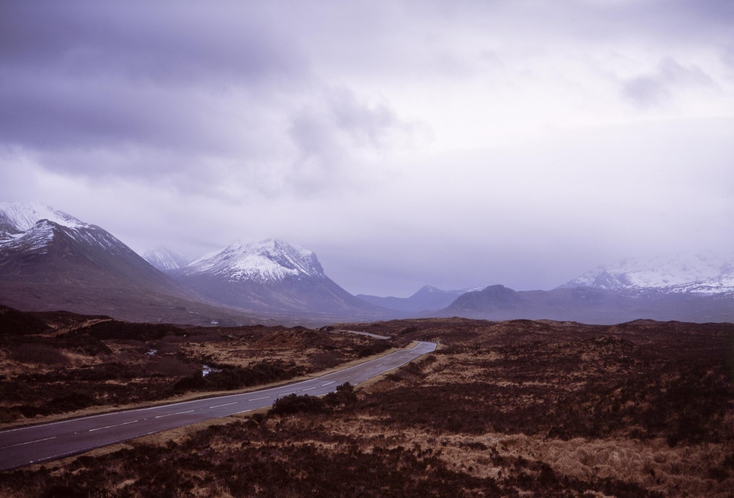 A winding road cuts through a vast, barren landscape with snow-capped mountains under a cloudy sky.