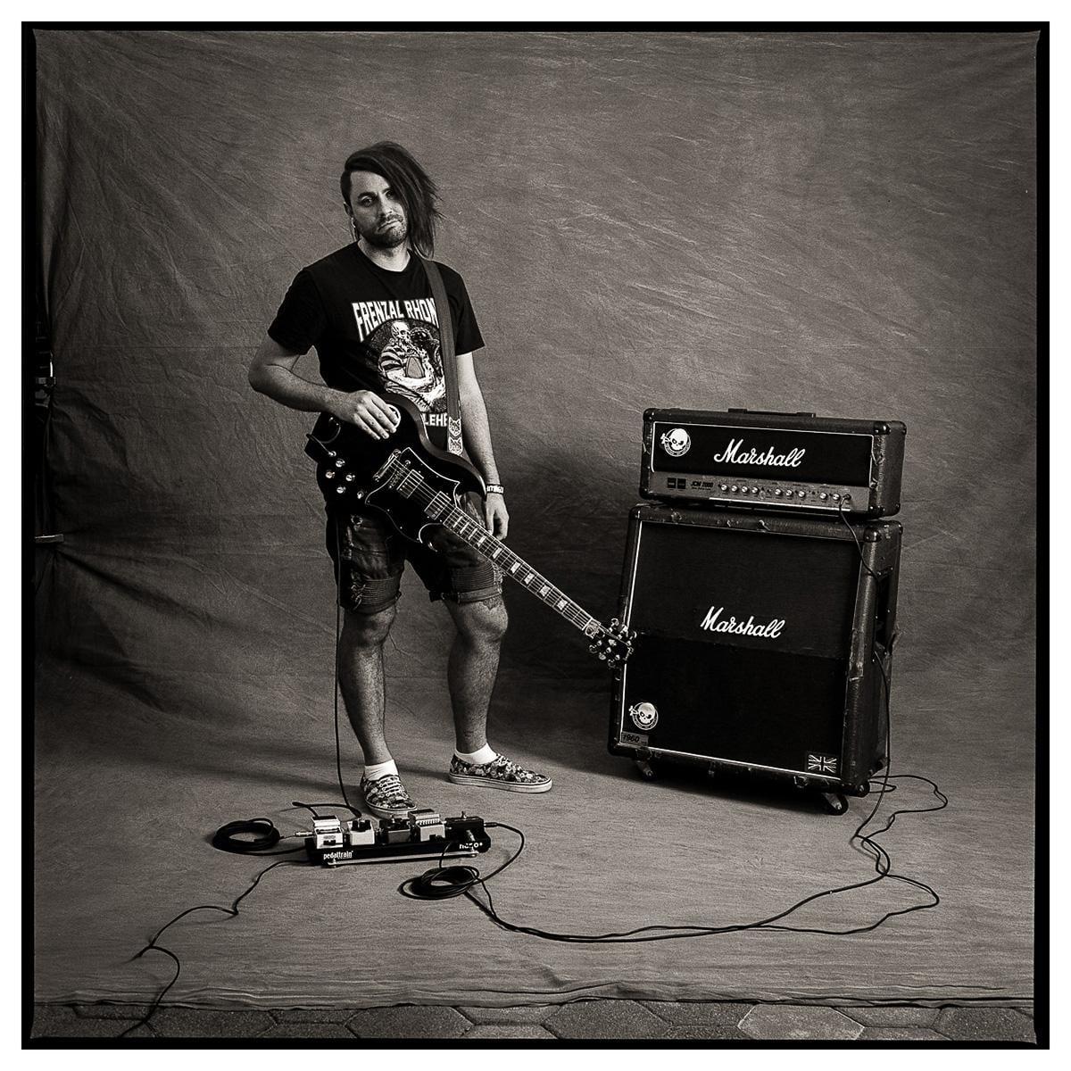 A man with long hair and a beard stands holding an electric guitar next to a stack of amplifiers and a pedalboard.