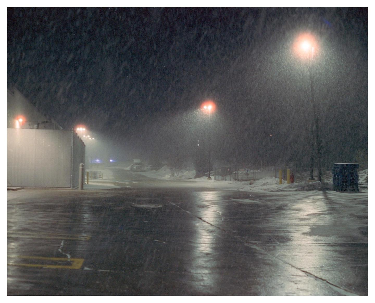 A snowy parking lot at night is illuminated by orange streetlights.