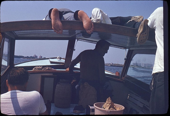 Three men are on a boat, with two men visible from the waist down relaxing on the roof of the boat, and one man in the cabin.