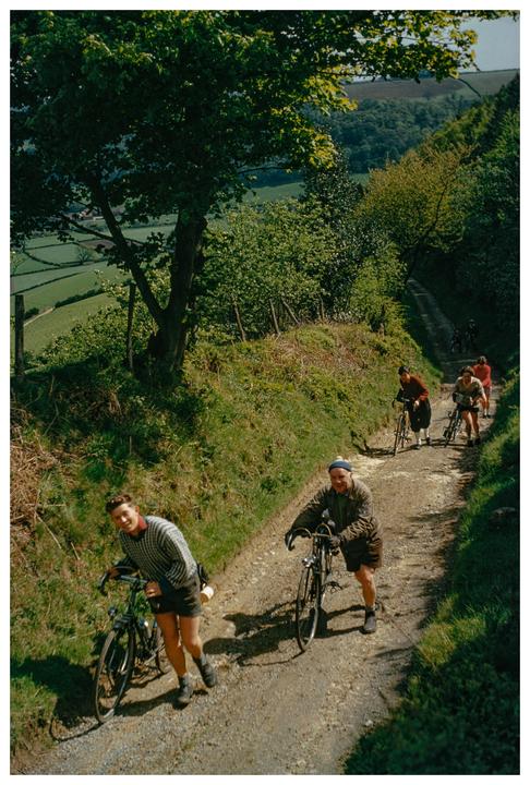 A group of cyclists push their bicycles up a steep, winding dirt path through a lush green landscape.