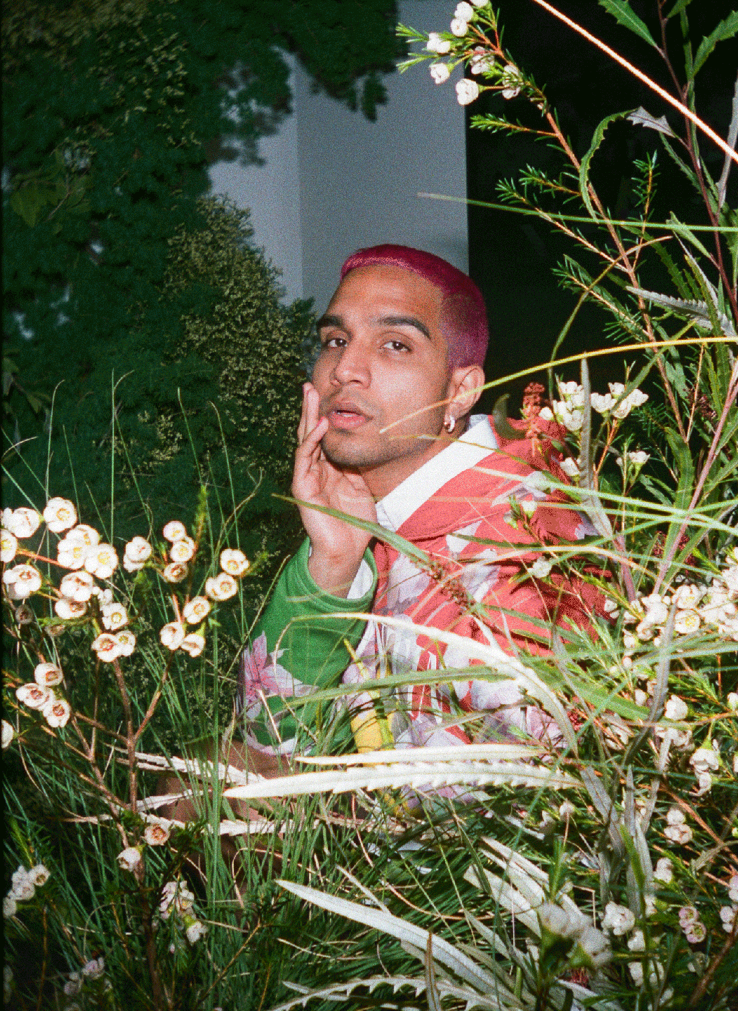 A man with pink hair and a colorful jacket is partially obscured by plants and flowers.