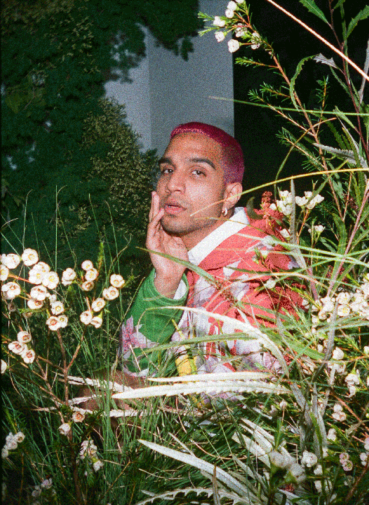 A man with pink hair and a colorful jacket is partially obscured by plants and flowers.