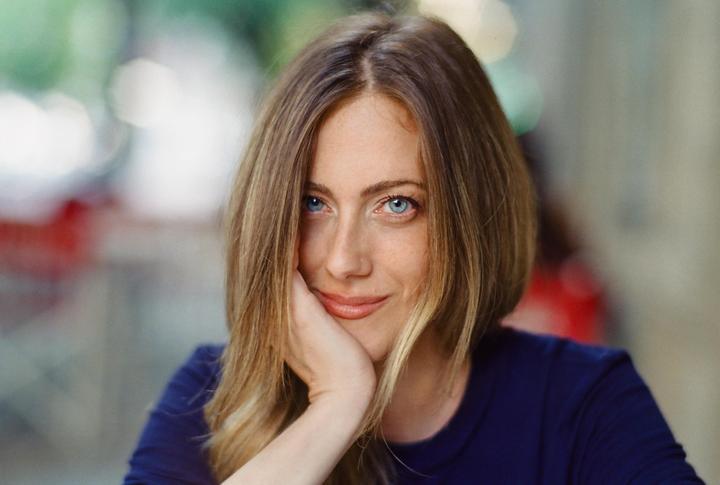 A woman with blue eyes and long hair rests her chin on her hand and smiles at the viewer.
