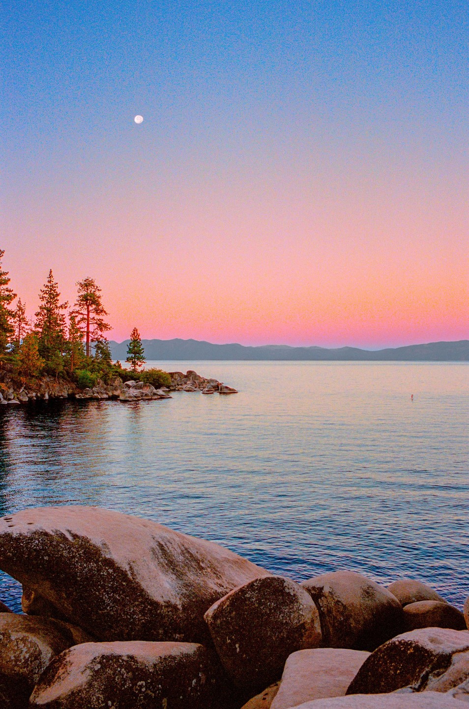 A full moon hangs in a colorful sky over a lake with a rocky shore and evergreen trees.