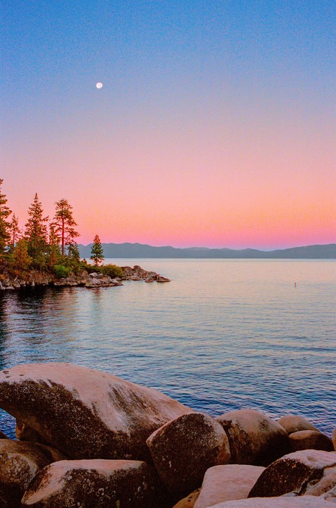 A full moon hangs in a colorful sky over a lake with a rocky shore and evergreen trees.