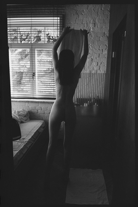 A woman stands nude with her back to the camera, holding a towel above her head in a room with a window and a bed.