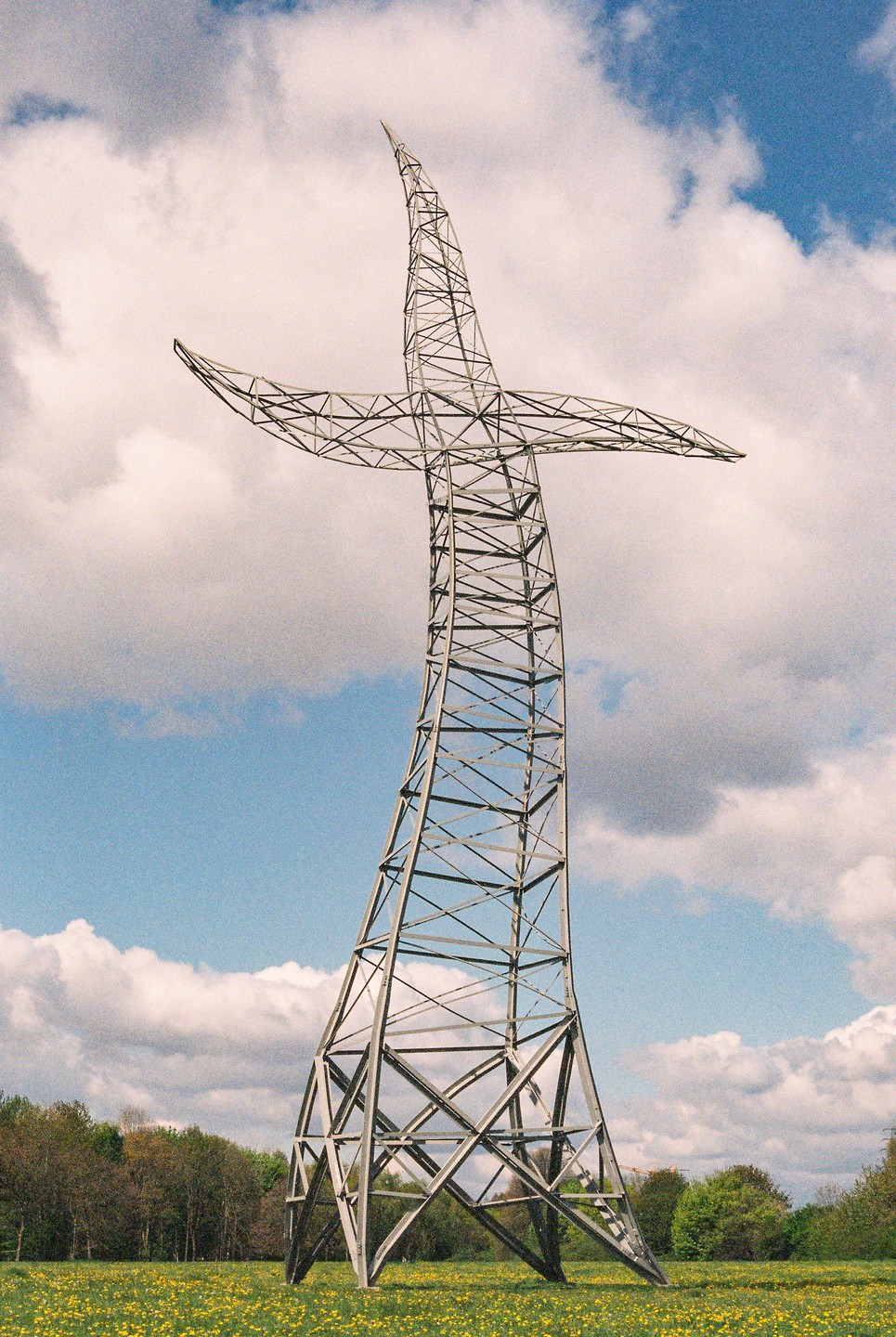 A large metal sculpture in the shape of a cross stands in a field of yellow flowers under a blue sky with white clouds.
