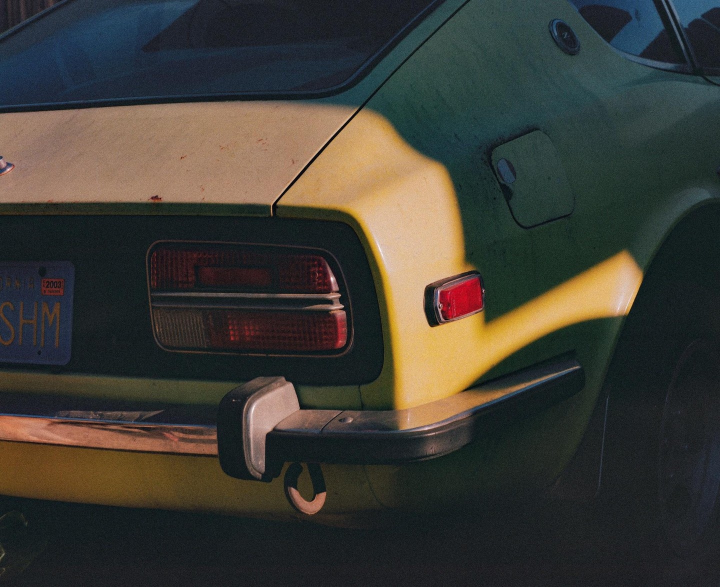 The rear of a green car with a California license plate is illuminated by a diagonal beam of sunlight.