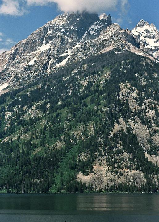 A large mountain with snow-capped peaks and green trees rises above a body of water under a blue sky with clouds.