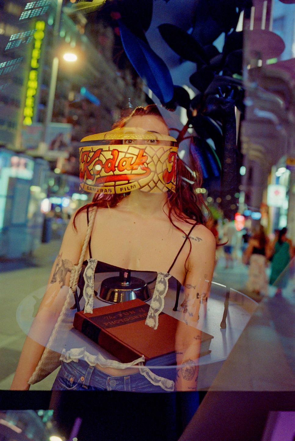 A woman with a Kodak film box on her face stands on a street at night.