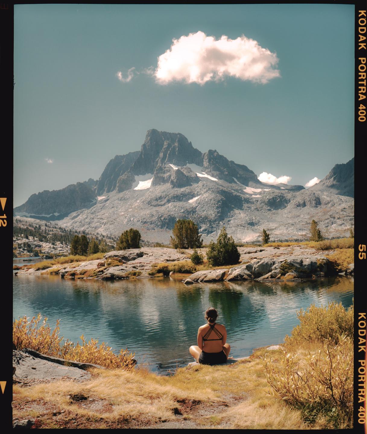 A woman sits by a lake with mountains in the background.