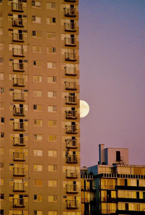 A full moon is visible behind a tall apartment building with many balconies.