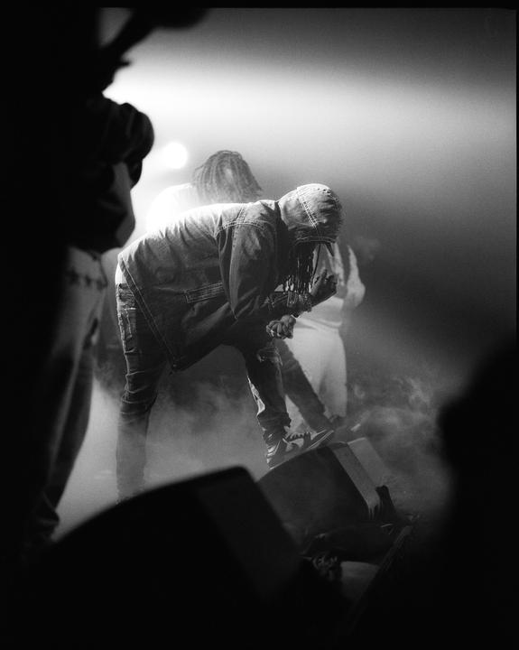 A man in a hooded denim jacket bends over on a stage with smoke and bright lights.