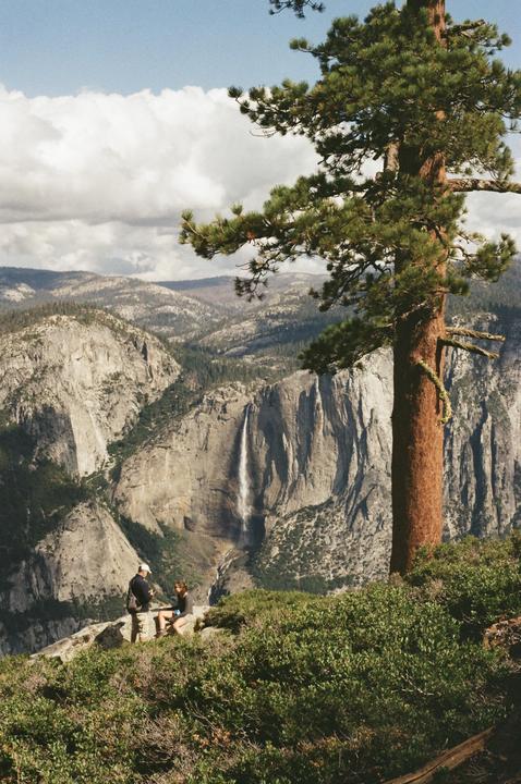 A couple sits on a rocky outcrop overlooking a valley with a waterfall and mountains.