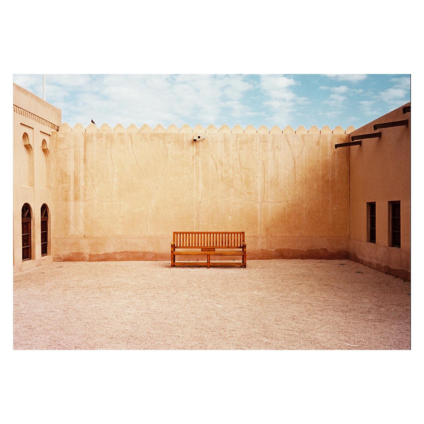 A wooden bench sits in a gravel courtyard surrounded by light brown walls with arched windows and a crenellated top.