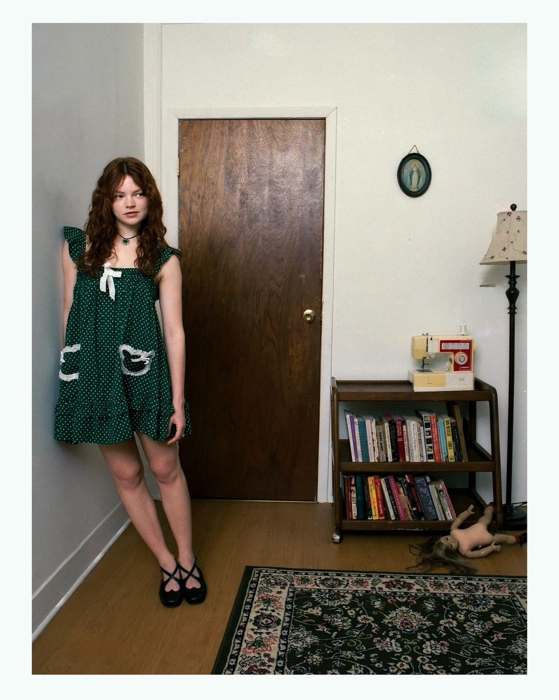 A woman in a green polka dot dress stands in a corner next to a wooden door, with a bookshelf, sewing machine, and doll visible.