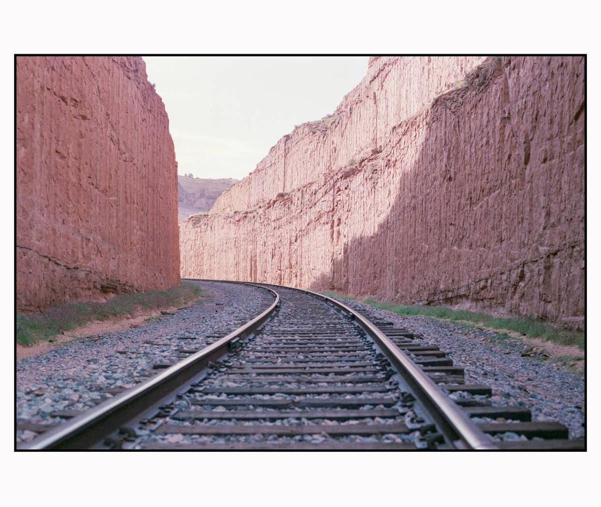 A set of train tracks curves through a narrow canyon with tall, reddish rock walls.
