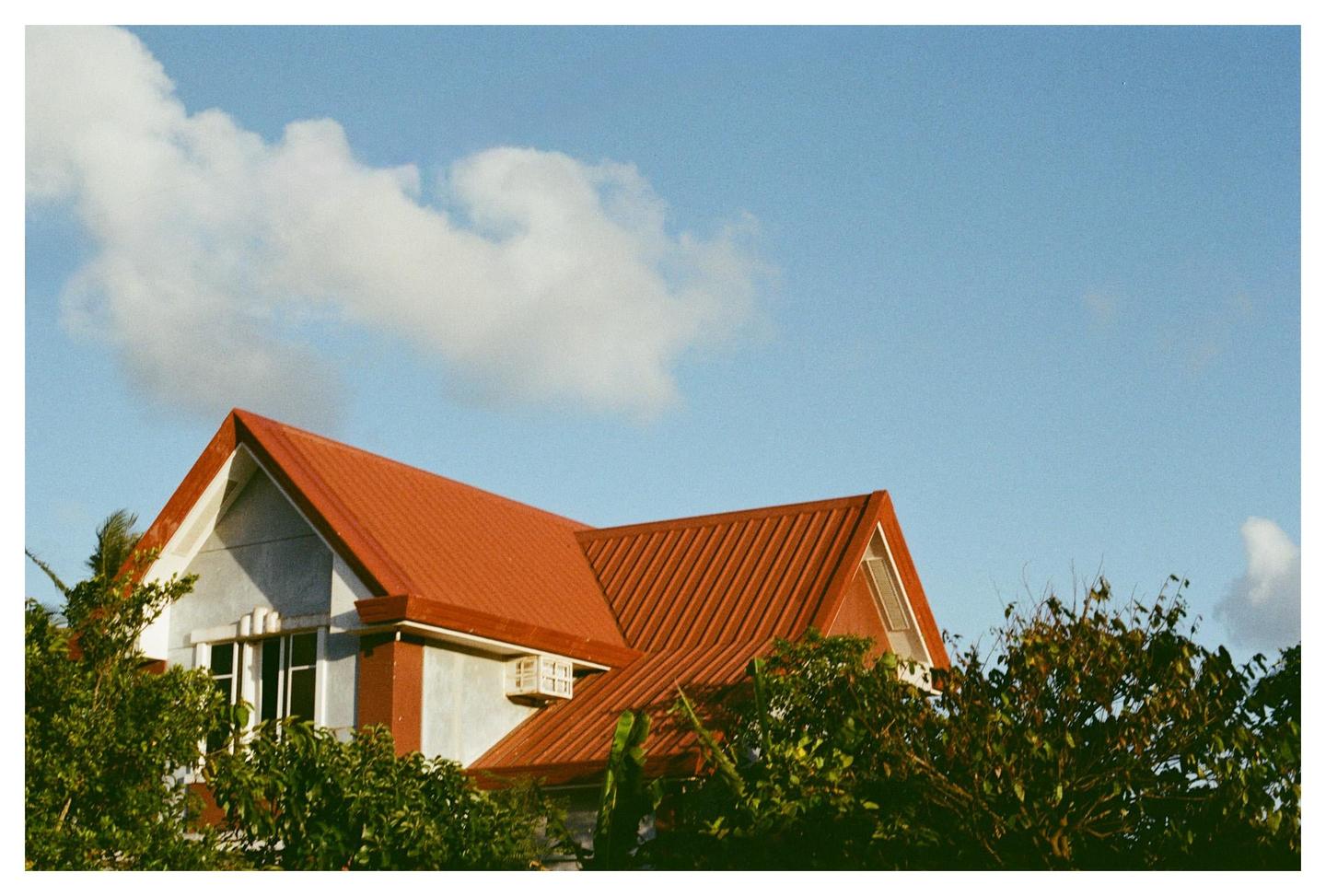 A house with a red roof is partially obscured by green foliage under a blue sky with a white cloud.