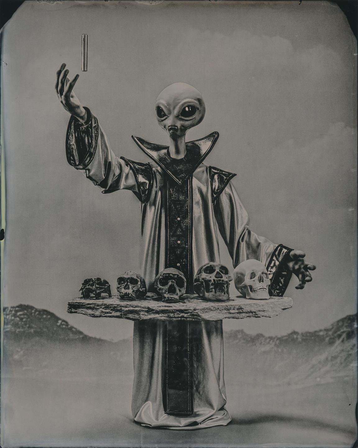 An alien in a robe holds a floating rectangular object in one hand and a tray of skulls in the other.
