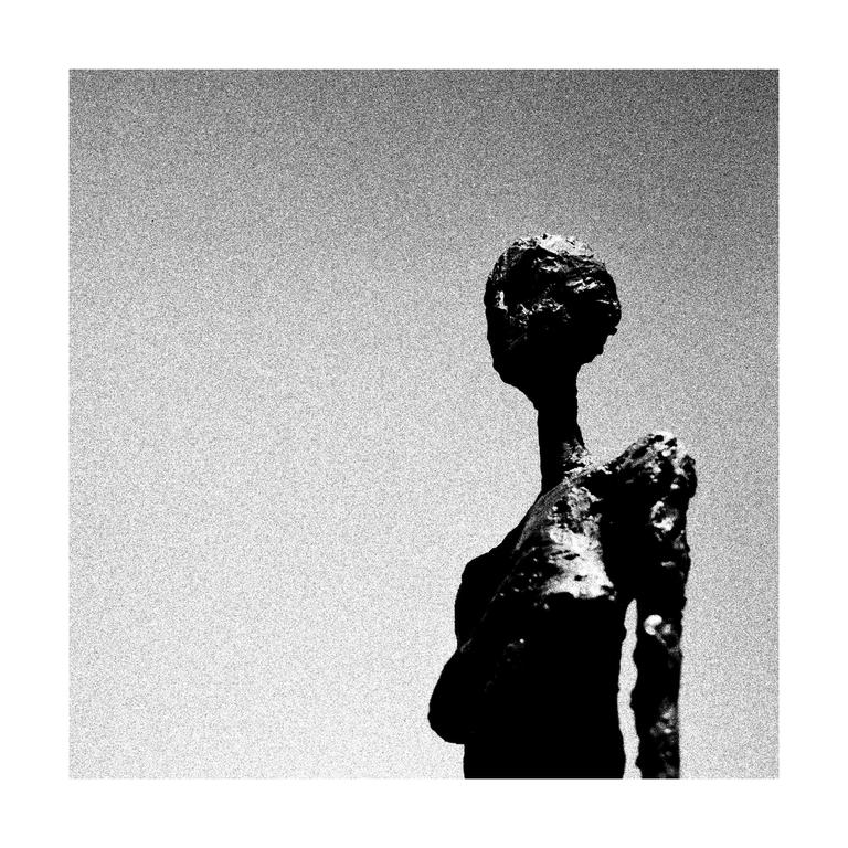 A dark, textured sculpture of a figure is silhouetted against a bright, grainy sky.