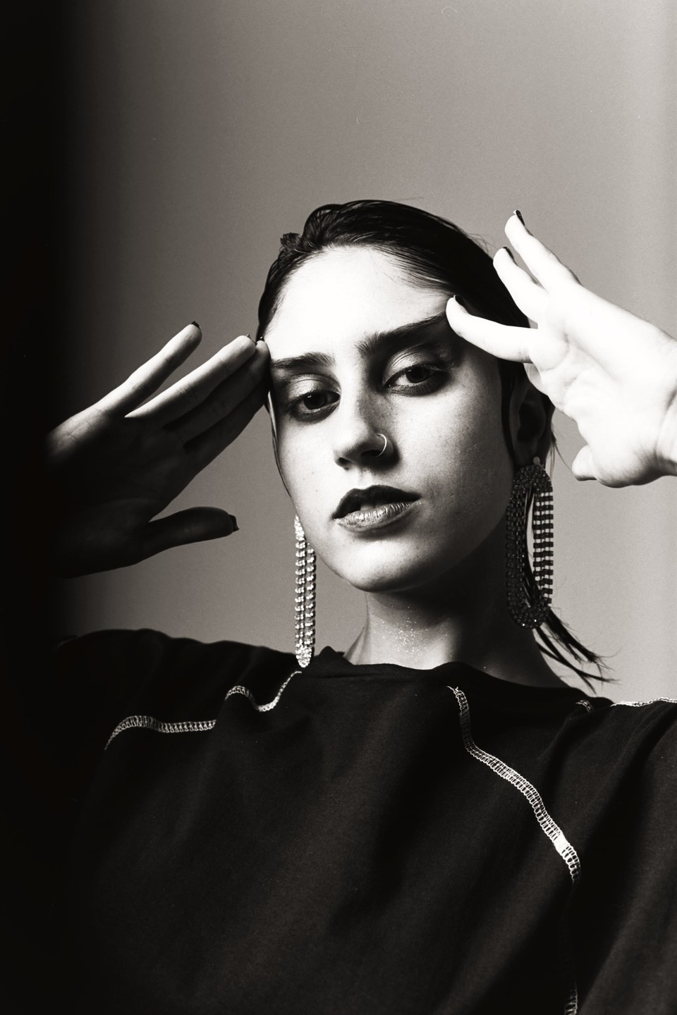A woman with dark hair and dramatic eye makeup poses with her hands near her temples, wearing large dangling earrings and a dark top with visible stitching.