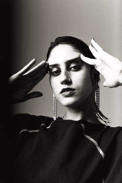 A woman with dark hair and dramatic eye makeup poses with her hands near her temples, wearing large dangling earrings and a dark top with visible stitching.