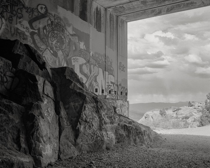 A concrete underpass covered in graffiti stands next to a rocky outcrop, with a view of distant mountains under a cloudy sky.