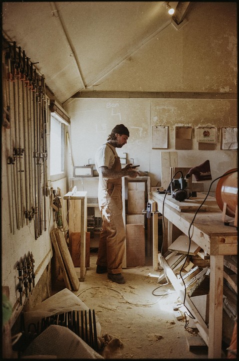 A man in overalls stands in a workshop, looking at a small object in his hands.