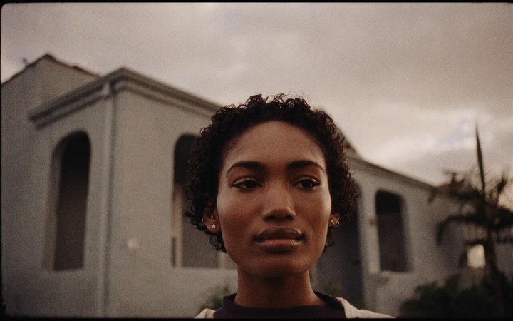 A woman with short curly hair looks directly at the viewer, with a building and palm tree in the background under a cloudy sky.