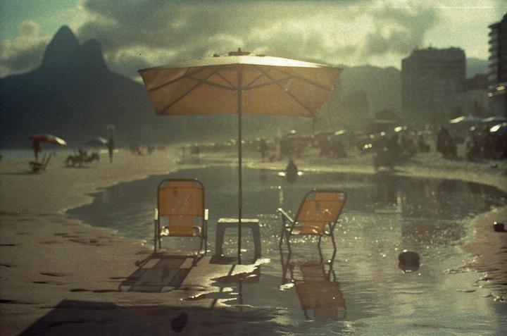 An orange beach umbrella and two chairs cast long shadows on a wet beach with mountains in the background.