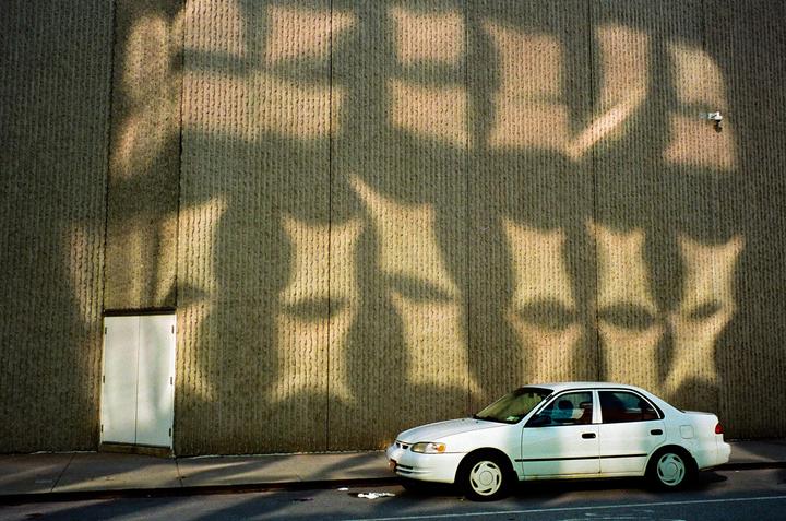 A white sedan is parked on a street next to a textured wall with abstract shadows.