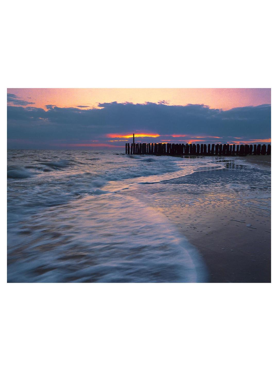 Waves crash on a beach next to a wooden pier at sunset.