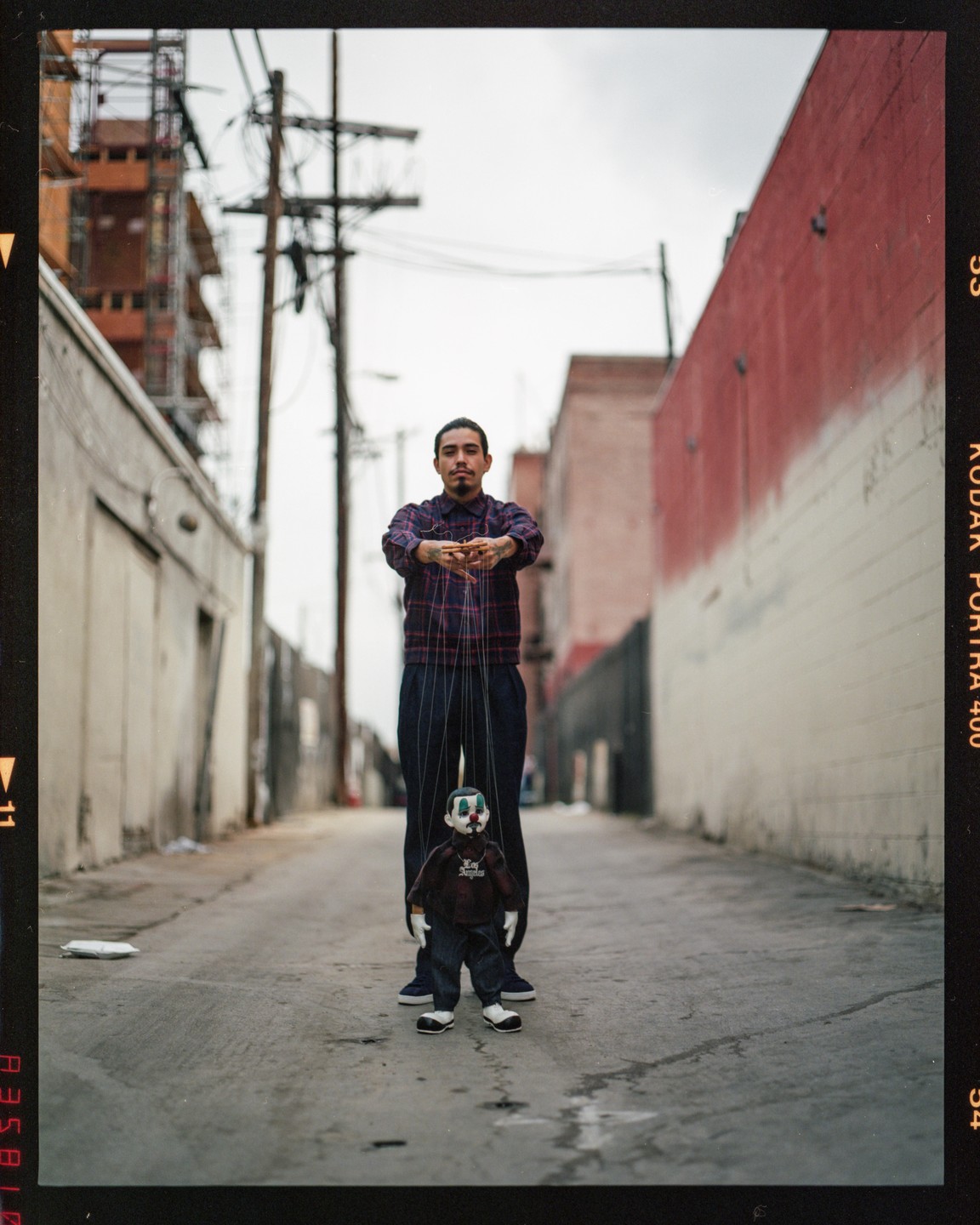 A man stands in an alley holding the strings of a puppet dressed in jeans and a hoodie.