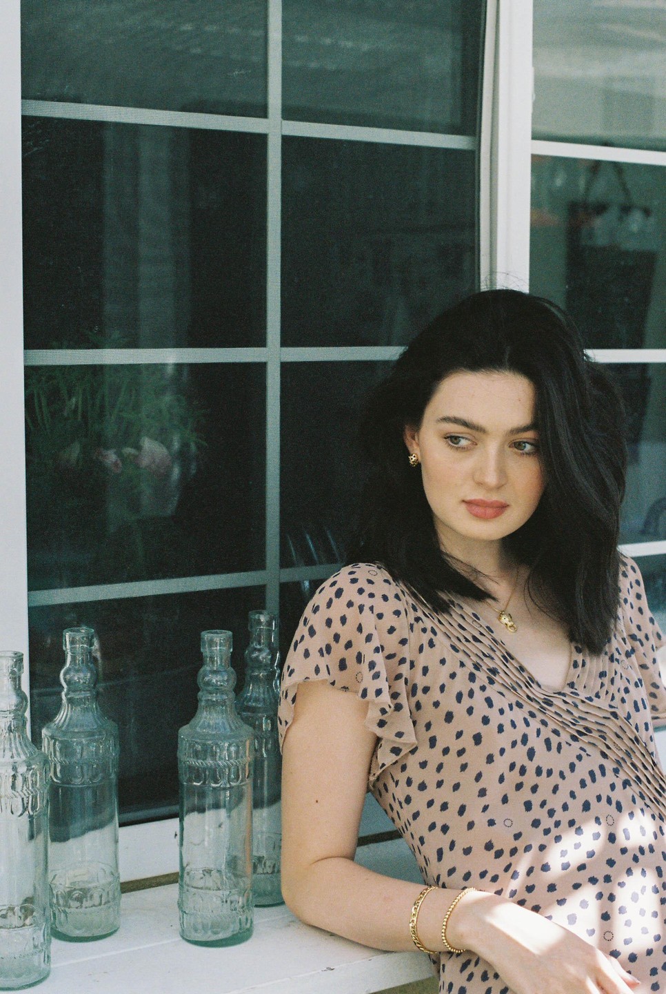 A woman in a spotted dress leans against a window with several glass bottles on the ledge.
