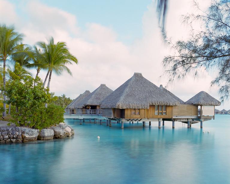 Overwater bungalows with thatched roofs stand in clear blue water next to a tropical island.