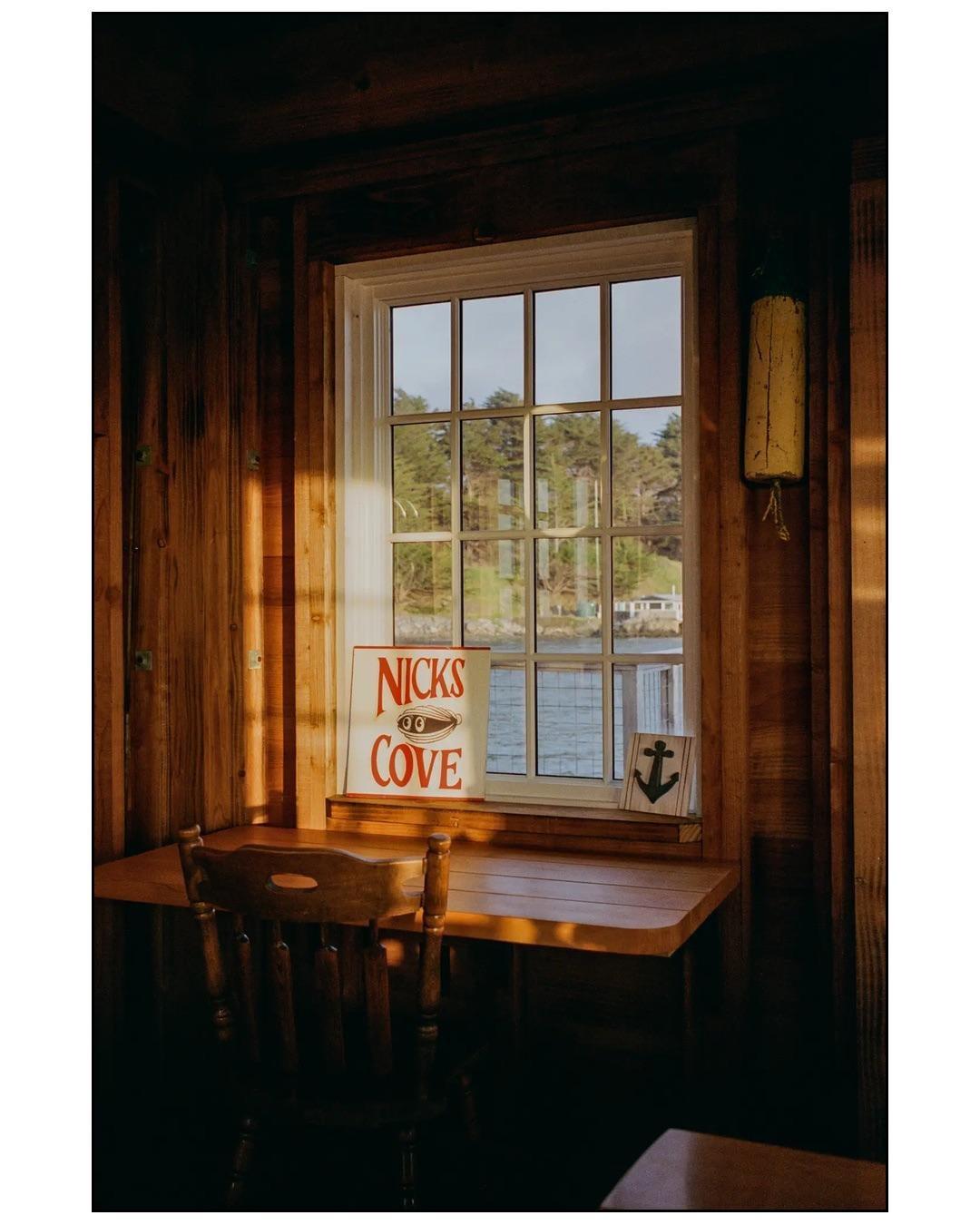 A wooden desk and chair are positioned in front of a window overlooking a body of water and trees.
