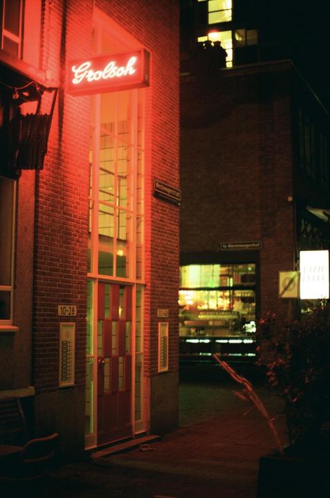 A red neon sign for Grolsch is mounted on a brick building with a tall window and red door at night.