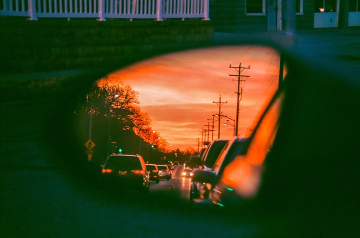 A street with cars and utility poles is reflected in a car's rearview mirror at sunset.