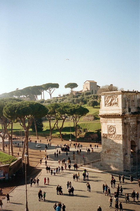 A large crowd of people gathers in a paved square next to a large stone archway, with a grassy hill and trees in the background.