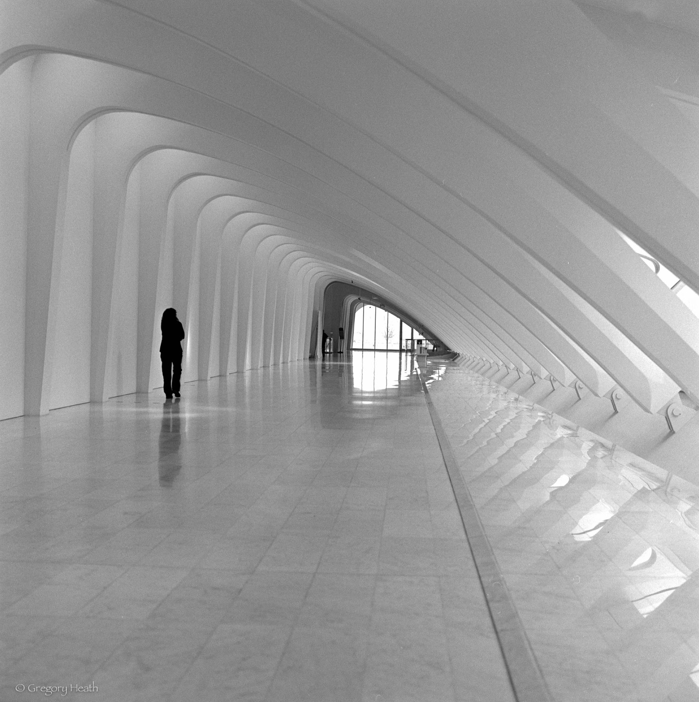 A lone woman stands in a long, modern, arched hallway with a bright window at the far end.